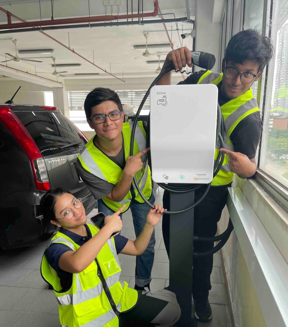 Three ITE students in safety vests pointing to a Schneider EVlink AC charger on a pedestal in a car park