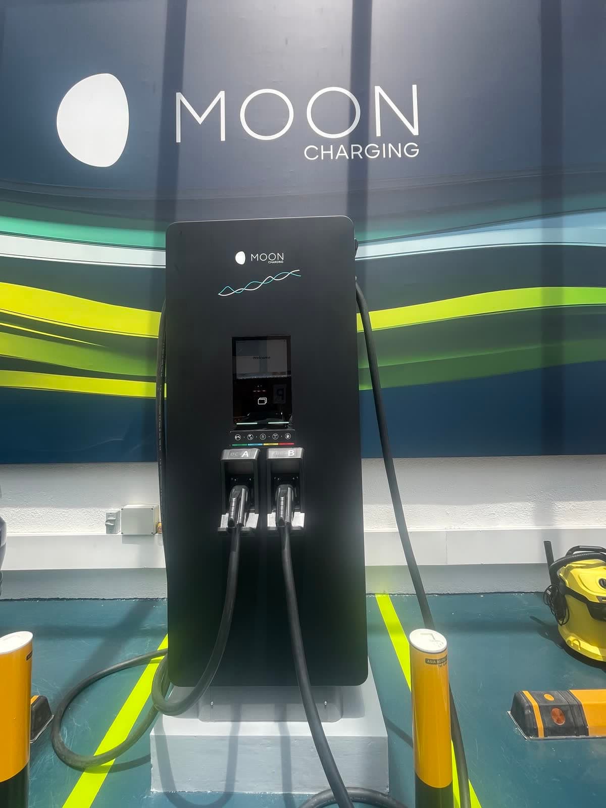 Dual-cable DC fast charger on a plinth between yellow bollards in a car park EV bay