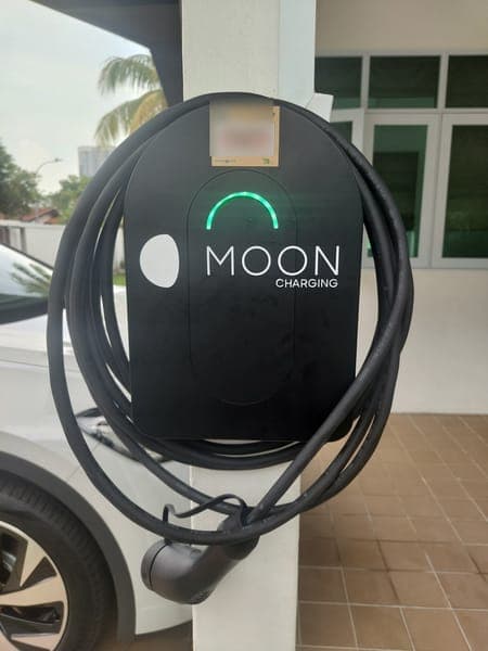 Black Moon Charging wallbox with a green light and coiled cable on a car porch pillar, a white car parked beside it