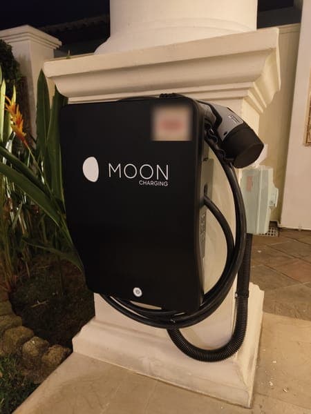 Moon Charging wallbox on a white porch pillar at night, beside tropical plants