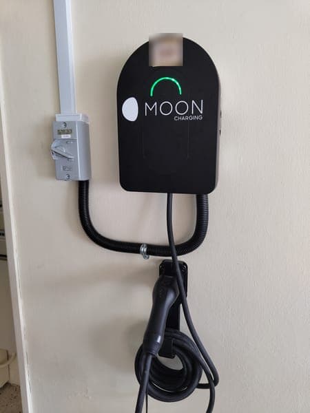 Moon Charging wallbox with a holstered plug and a conduit loop on a cream wall