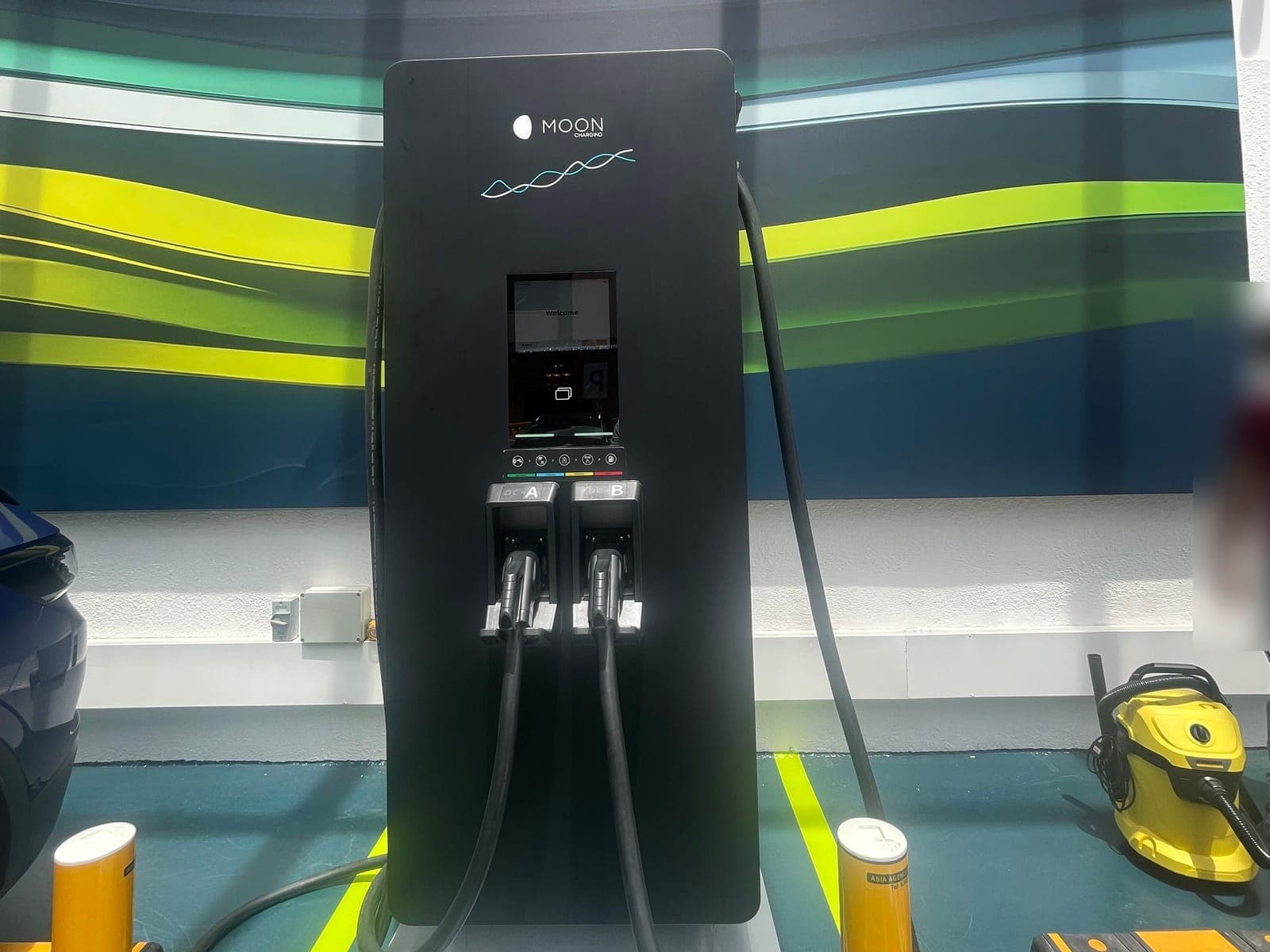 Dual-gun DC fast charger with a touchscreen and two holstered DC connectors in a covered car park bay
