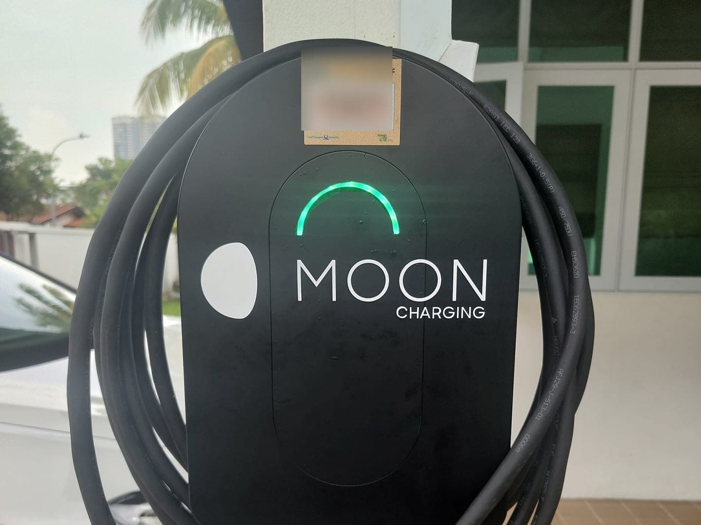 Black Moon Charging wallbox with a green status light and coiled cable on a car porch pillar, with a white car parked alongside