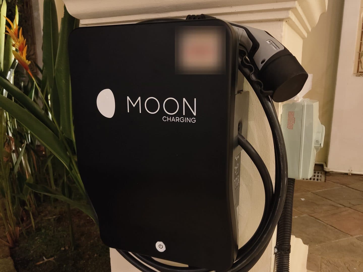 Black Moon Charging wallbox with its Type 2 cable and isolator on a white car porch pillar at night, beside tropical plants